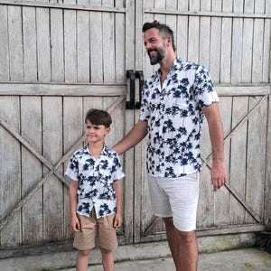 K&ouml;nnte beinhalten: Ein Vater und sein Sohn tragen passende wei&szlig;e Hemden mit kurzen &Auml;rmeln und einem blauen Palmenprint. Die Hemden haben einen Knopfkrag und kurze &Auml;rmel. Der Vater tr&auml;gt wei&szlig;e Shorts und der Sohn tr&auml;gt Khaki-Shorts. Beide l&auml;cheln und stehen vor einer Holzt&uuml;r.