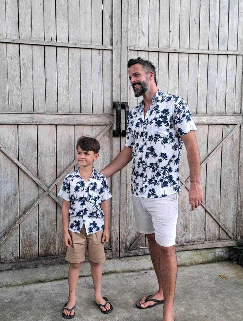 K&ouml;nnte beinhalten: Ein Vater und sein Sohn tragen passende wei&szlig;e Hemden mit kurzen &Auml;rmeln und einem blauen Palmenprint. Die Hemden haben einen Knopfkrag und kurze &Auml;rmel. Der Vater tr&auml;gt wei&szlig;e Shorts und der Sohn tr&auml;gt Khaki-Shorts. Beide l&auml;cheln und stehen vor einer Holzt&uuml;r.