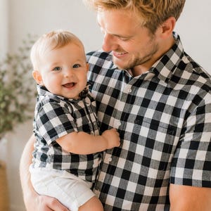 Könnte beinhalten: Ein Vater und ein Kind sind in einer herzlichen Umarmung zu sehen, beide tragen passende schwarz-weiß karierte Hemden. Das Kind trägt auch weiße Shorts. Der Vater hält das Kind, beide lächeln in die Kamera.