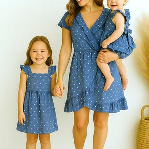 May include: A family of three wearing matching blue dresses with white polka dots. The dresses have ruffled hems and sleeves. The mother is holding a baby, and a young girl is holding her hand. The dresses are perfect for a family photo.