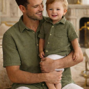 May include: A man and a toddler, both in olive green short-sleeved shirts and white trousers. The man is holding the child and smiling. The child is looking at the camera and smiling. The setting appears to be indoors.