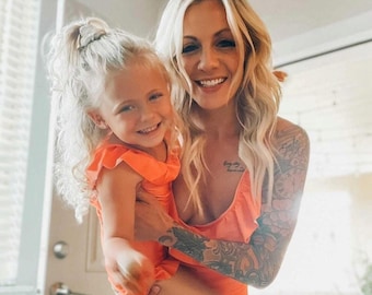 May include: A woman and a young girl are wearing matching orange ruffled swimsuits. The woman has blonde hair and a tattoo on her arm. The girl has blonde hair and is smiling.