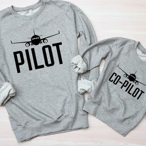 Könnte beinhalten: Zwei graue Sweatshirts mit einem schwarzen Flugzeug-Motiv und Text. Das größere Sweatshirt sagt "PILOT" und das kleinere Sweatshirt sagt "CO-PILOT".