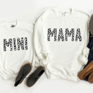 Puede incluir: Dos sudaderas blancas con contornos negros de las palabras "MINI" y "MAMA" en un estampado de leopardo.