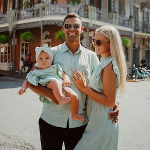 Puede incluir: Una familia de tres posa al aire libre, todos con conjuntos verde claro a juego. El padre sostiene al beb&eacute;, que lleva una diadema a juego. La madre lleva un vestido. Sonr&iacute;en frente a un edificio con un balc&oacute;n de hierro forjado.