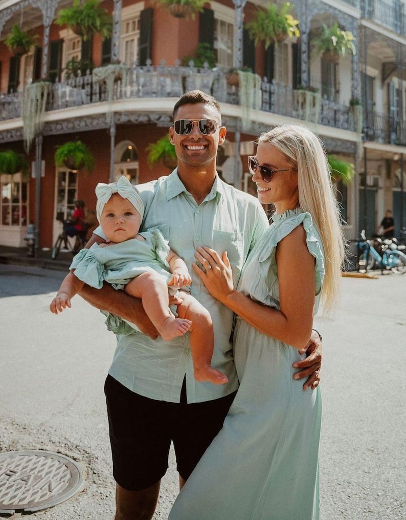 Puede incluir: Una familia de tres posa al aire libre, todos con conjuntos verde claro a juego. El padre sostiene al beb&eacute;, que lleva una diadema a juego. La madre lleva un vestido. Sonr&iacute;en frente a un edificio con un balc&oacute;n de hierro forjado.