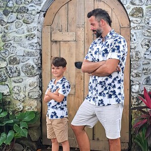 K&ouml;nnte beinhalten: Ein Vater und sein Sohn tragen passende wei&szlig;e Hemden mit kurzen &Auml;rmeln und einem blauen Palmenbaumdruck. Die Hemden haben einen Knopfkragenstil und kurze &Auml;rmel. Der Vater tr&auml;gt wei&szlig;e Shorts und der Sohn tr&auml;gt Khaki-Shorts.
