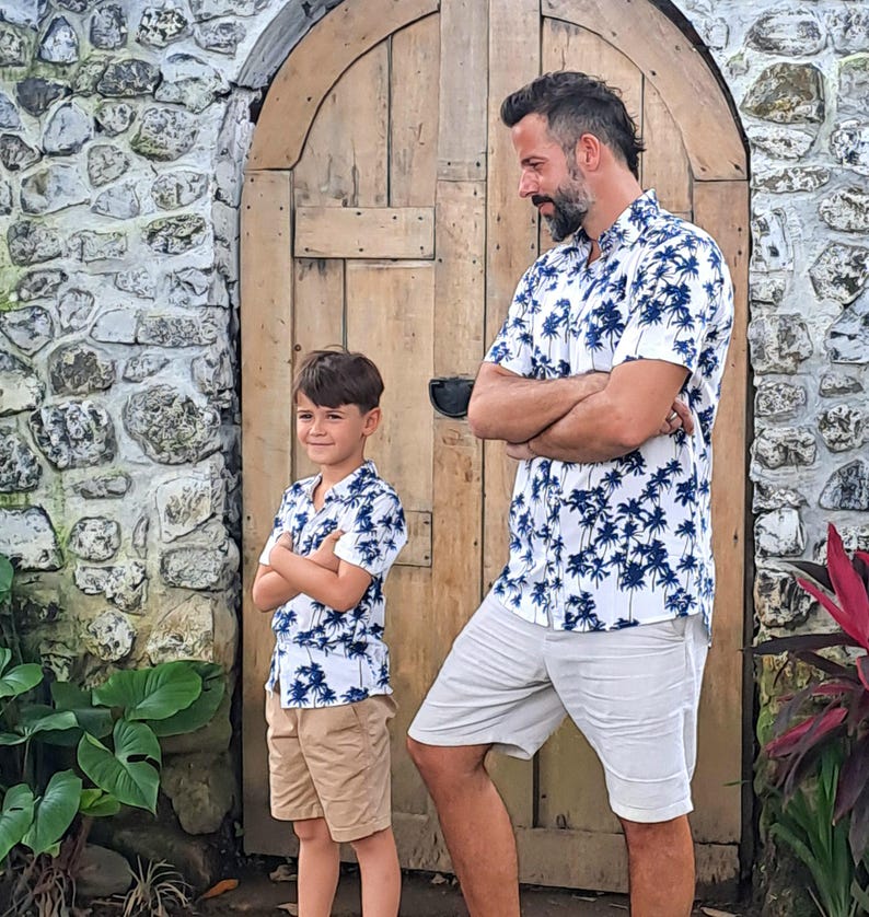 K&ouml;nnte beinhalten: Ein Vater und sein Sohn tragen passende wei&szlig;e Hemden mit kurzen &Auml;rmeln und einem blauen Palmenbaumdruck. Die Hemden haben einen Knopfkragenstil und kurze &Auml;rmel. Der Vater tr&auml;gt wei&szlig;e Shorts und der Sohn tr&auml;gt Khaki-Shorts.