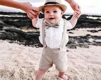 May include: A baby boy wearing a white shirt, beige linen shorts, a brown bow tie, and a brown fedora hat. The baby is standing on a sandy beach.