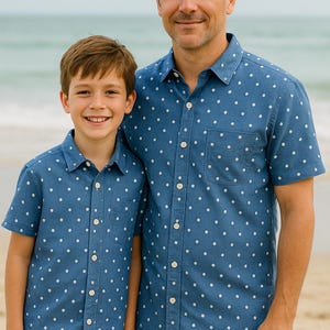 May include: Two people wearing matching blue button-down shirts with white polka dots and khaki shorts. The shirts have a chest pocket. They are standing on a sandy beach with the ocean in the background.