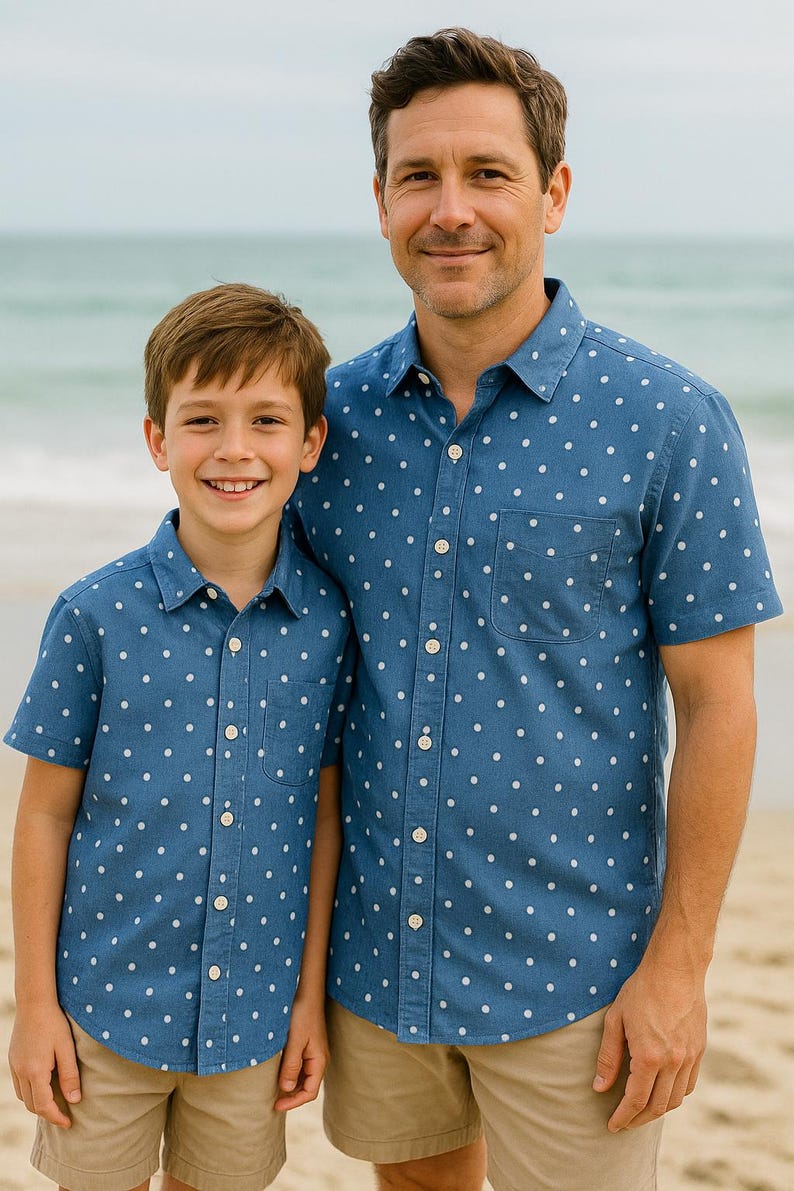 May include: Two people wearing matching blue button-down shirts with white polka dots and khaki shorts. The shirts have a chest pocket. They are standing on a sandy beach with the ocean in the background.