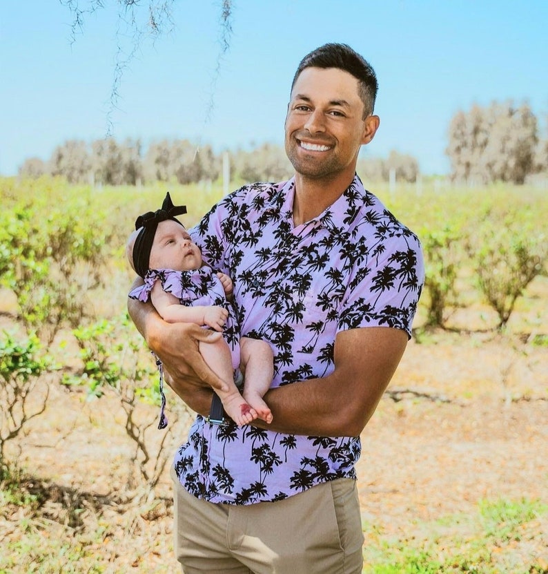 K&ouml;nnte beinhalten: Ein Mann in einem lila und schwarzen Hemd mit Palmenprint h&auml;lt ein kleines M&auml;dchen in einem wei&szlig;en Kleid mit einem schwarzen Schleifenhaarband. Das Baby schaut in die Kamera und der Mann l&auml;chelt.