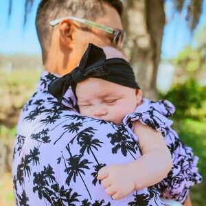 K&ouml;nnte beinhalten: Ein Mann in einem pink-schwarzen Hemd mit Palmenprint h&auml;lt ein schlafendes Baby, das ein rosa-wei&szlig;es Kleid mit einem schwarzen Schleifenhaarband tr&auml;gt.