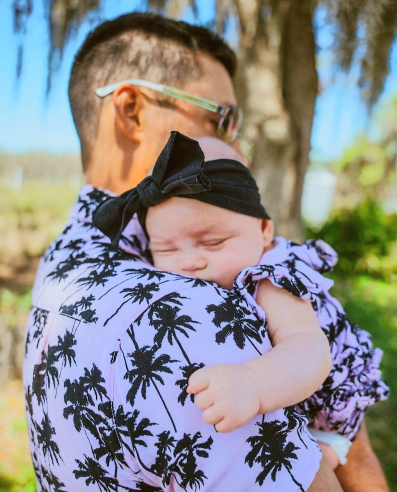 K&ouml;nnte beinhalten: Ein Mann in einem pink-schwarzen Hemd mit Palmenprint h&auml;lt ein schlafendes Baby, das ein rosa-wei&szlig;es Kleid mit einem schwarzen Schleifenhaarband tr&auml;gt.