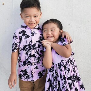 K&ouml;nnte beinhalten: Zwei Kinder tragen passende lila Hemden mit einem schwarzen Palmen-Druck. Der Junge tr&auml;gt eine braune Hose und das M&auml;dchen tr&auml;gt ein lila Kleid. Sie l&auml;cheln und schauen in die Kamera.