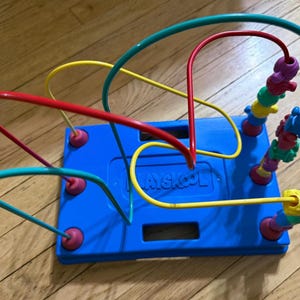 May include: A blue Playskool bead maze toy with colorful wire loops and multi-colored beads. The base is rectangular with the word "Playskool" printed on it. The wires are red, yellow, and teal.