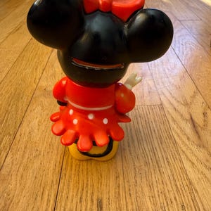 Op de afbeelding: Een Minnie Mouse-beeldje, met een zwarte kop, grote oren en een rode strik. Het beeldje draagt een rode jurk met witte stippen en een gele basis. Dit is een verzamelobject.