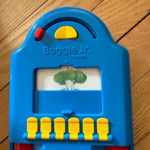 May include: A blue Boggle Jr. Travel game with yellow buttons and accents. The game features a picture of a tree and the text "Boggle Jr. Travel". The game has red knobs on the sides.