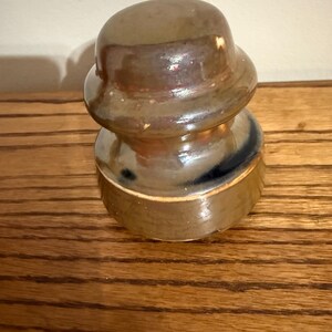 May include: A vintage glass electrical insulator with a smooth, rounded top and a wider, cylindrical base. The object features a glossy, multi-toned finish with blended shades of brown, tan, and dark blue, reflecting light across its curved surface.