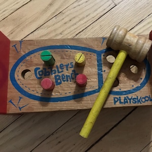 May include: A vintage wooden "Cobblers Bench" toy by Playskool. The light brown base has a blue oval design and colourful pegs. A wooden mallet with a yellow handle lies on the bench. Red end pieces complete the toy.