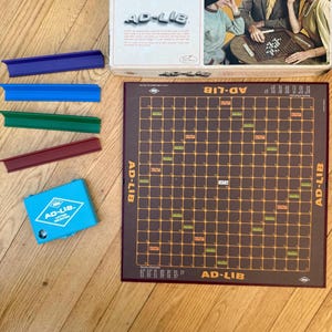 May include: A vintage AD-LIB board game set. The game includes a brown game board with a grid pattern, letter holders in blue, green, and red, and a blue letter selector. The box features the game's name and an image of people playing.