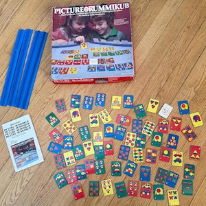 May include: A vintage Picture Rummikub game set. The box features children playing the game. The set includes blue plastic holders, a booklet, and numerous colorful tiles with various pictures and numbers. The game is on a wooden surface.