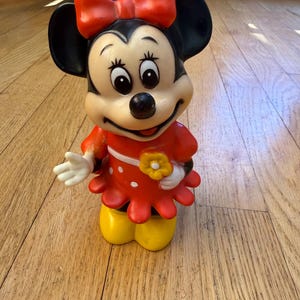 Op de afbeelding: Een vintage Minnie Mouse beeldje. Het figuur draagt een rode jurk met witte stippen, een rode strik en gele schoenen. Het beeldje houdt een gele bloem vast en heeft zwarte oren.