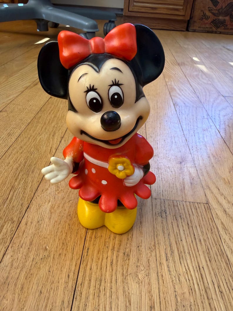 Op de afbeelding: Een vintage Minnie Mouse beeldje. Het figuur draagt een rode jurk met witte stippen, een rode strik en gele schoenen. Het beeldje houdt een gele bloem vast en heeft zwarte oren.