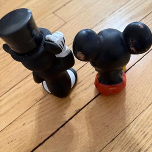 May include: Two vintage Mickey Mouse figurines. One figure is dressed in a black tuxedo with a top hat and white gloves. The other figure is black with red shorts and yellow shoes. Both figures are on a wooden surface.