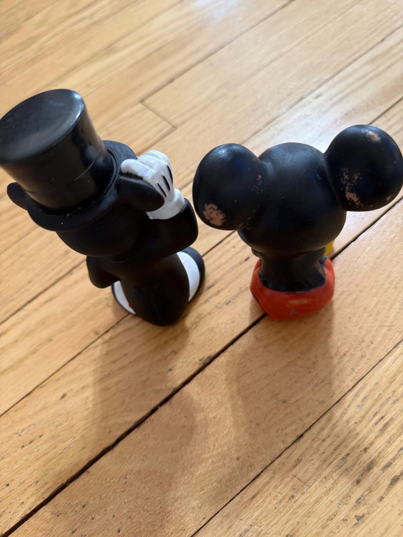 May include: Two vintage Mickey Mouse figurines. One figure is dressed in a black tuxedo with a top hat and white gloves. The other figure is black with red shorts and yellow shoes. Both figures are on a wooden surface.