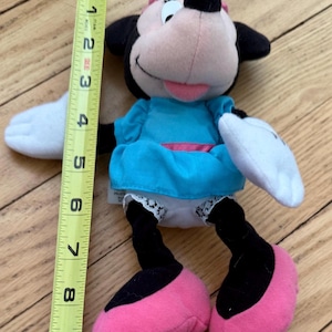May include: A Minnie Mouse soft toy, approximately 23 cm tall, with a pink bow, black ears, and a light blue dress. The toy has pink shoes, white gloves, and black legs. The plush is on a wooden surface.
