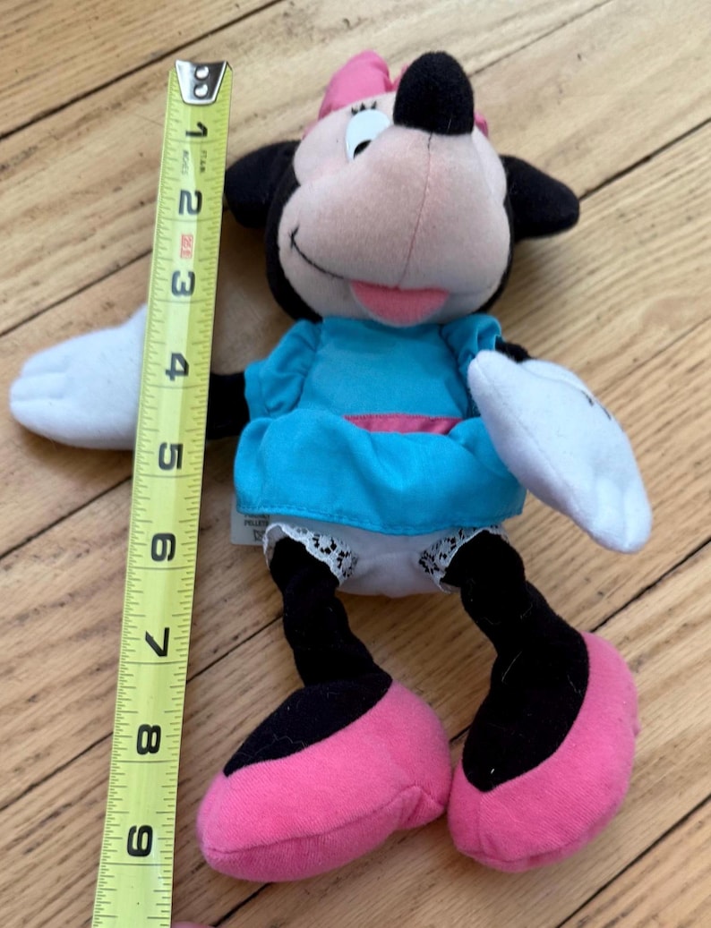 May include: A Minnie Mouse soft toy, approximately 23 cm tall, with a pink bow, black ears, and a light blue dress. The toy has pink shoes, white gloves, and black legs. The plush is on a wooden surface.