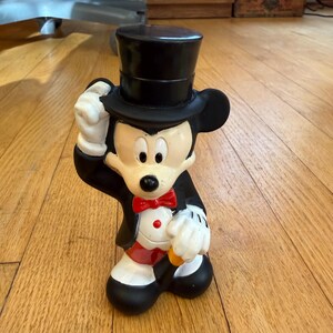 May include: A ceramic Mickey Mouse figurine in a tuxedo and top hat. The black and white figure has a red bow tie and is holding his hat. The figurine is standing on a wooden floor.