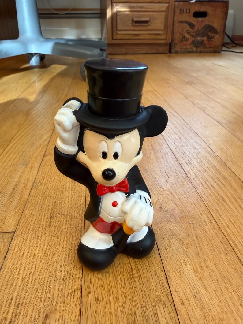 May include: A ceramic Mickey Mouse figurine in a tuxedo and top hat. The black and white figure has a red bow tie and is holding his hat. The figurine is standing on a wooden floor.