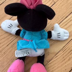May include: A Minnie Mouse soft toy, with a black head, large ears, and a pink bow. She is dressed in a blue dress with a pink belt, white gloves with black lines, and pink shoes. The toy is made of soft materials.