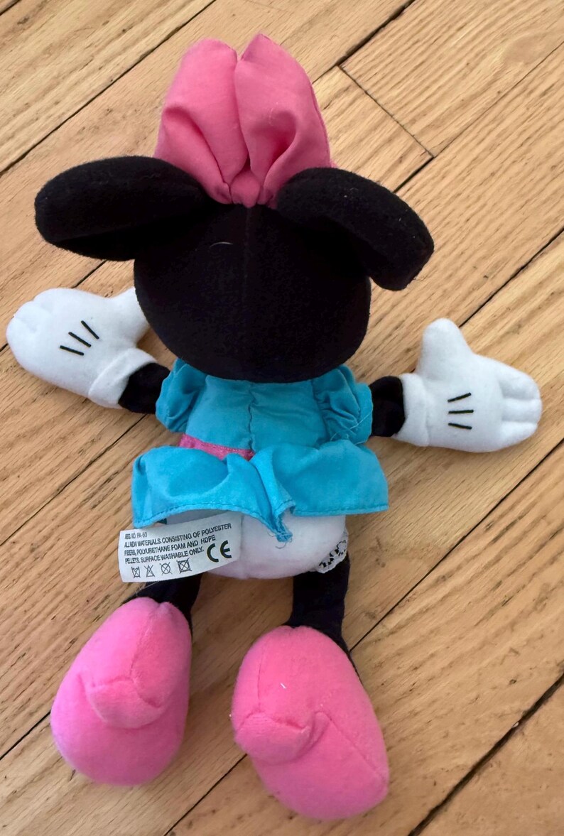 May include: A Minnie Mouse soft toy, with a black head, large ears, and a pink bow. She is dressed in a blue dress with a pink belt, white gloves with black lines, and pink shoes. The toy is made of soft materials.