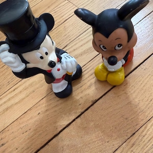 May include: Two Mickey Mouse figurines. One is in a tuxedo and top hat, the other is in a classic pose. Both are black, white, and yellow. The figurines are on a wooden surface.