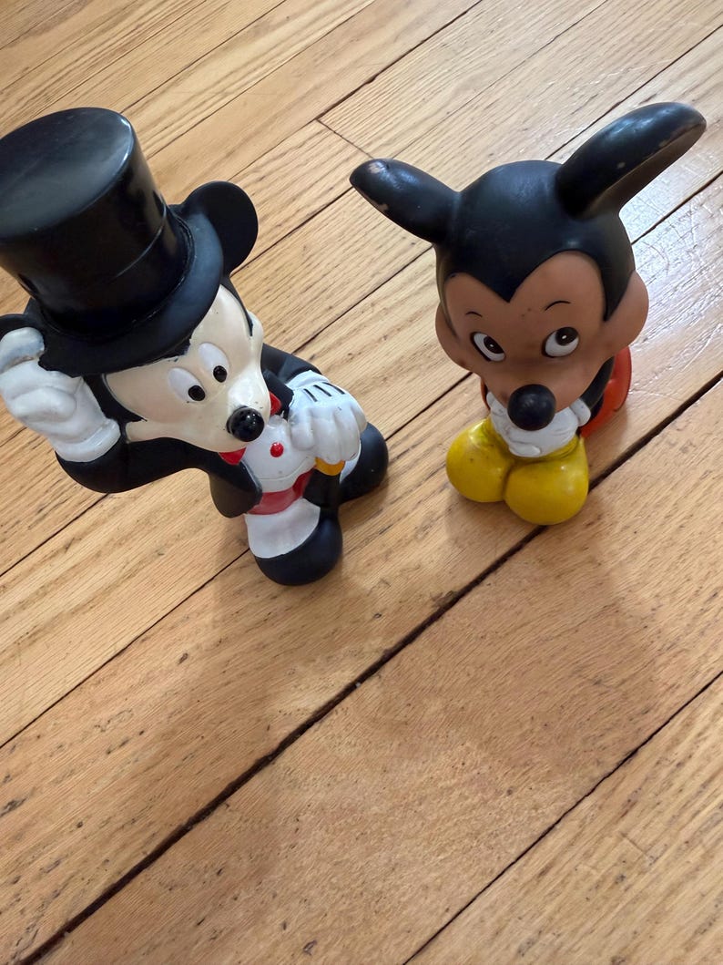 May include: Two Mickey Mouse figurines. One is in a tuxedo and top hat, the other is in a classic pose. Both are black, white, and yellow. The figurines are on a wooden surface.