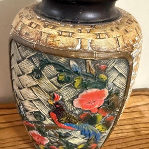 May include: A decorative vase featuring a dark brown rim and a textured, woven-look body. The surface displays a raised, colorful relief of a bird with red and blue plumage perched among vibrant pink and blue flowers against a basket-weave pattern.