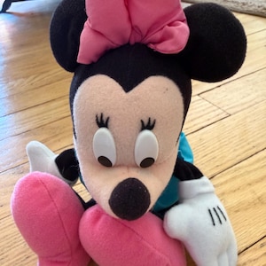 May include: A Minnie Mouse soft toy featuring a large pink bow, black ears, and a black nose. The doll has white gloves with black lines and pink shoes. The toy is positioned on a wooden floor.