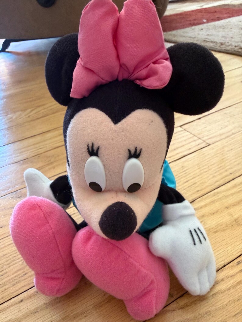 May include: A Minnie Mouse soft toy featuring a large pink bow, black ears, and a black nose. The doll has white gloves with black lines and pink shoes. The toy is positioned on a wooden floor.