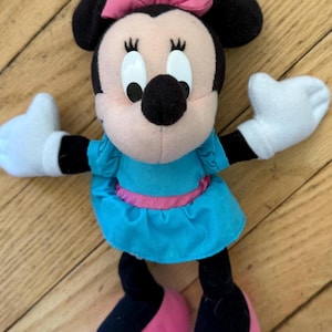 May include: A plush Minnie Mouse doll with black ears, a pink bow, and a light blue dress with a pink belt. The doll has white gloves and pink shoes. The doll is standing on a wooden surface.