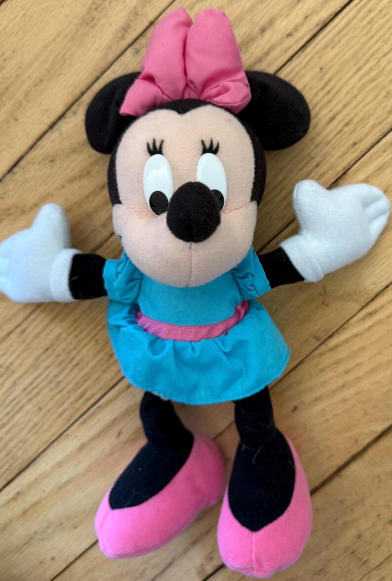 May include: A plush Minnie Mouse doll with black ears, a pink bow, and a light blue dress with a pink belt. The doll has white gloves and pink shoes. The doll is standing on a wooden surface.