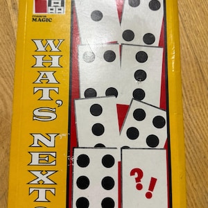 Op de afbeelding: Een rechthoekige gele speldoos met de titel WHAT'S NEXT? voorzien van een rood omrande afbeelding van witte speelkaarten met zwarte stippen. In de linkerbovenhoek staat een logo met de tekst means MAGIC. Eén kaart toont een rood vraagteken en uitroepteken.