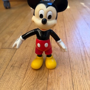 Op de afbeelding: Een Mickey Mouse-beeldje, met een zwart lichaam, oren en armen, en witte handschoenen. Het beeldje heeft een cr&egrave;mekleurig gezicht, een rode broek en gele schoenen. Het klassieke Disney-personage staat op een houten vloer.