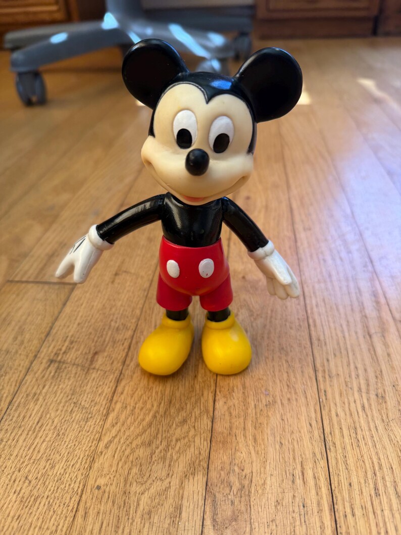 Op de afbeelding: Een Mickey Mouse-beeldje, met een zwart lichaam, oren en armen, en witte handschoenen. Het beeldje heeft een cr&egrave;mekleurig gezicht, een rode broek en gele schoenen. Het klassieke Disney-personage staat op een houten vloer.