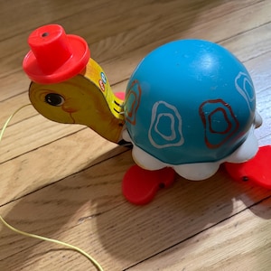 Puede incluir: Una tortuga de juguete vintage para tirar. La tortuga tiene una cabeza amarilla con un sombrero rojo y un caparazón azul decorado con detalles blancos y rojos. La tortuga tiene patas rojas y una cuerda amarilla.