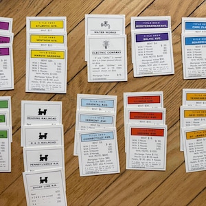 May include: A collection of vintage Monopoly property deed cards, featuring various street names and property details. The cards are in different colors, including purple, yellow, and green, and are arranged on a wooden surface.