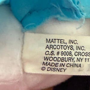 May include: A close-up of a white tag with black text. The tag reads "MATTEL, INC. ARCOTOYS, INC. C.S. #9008, CROSSWAYS PARK WEST, WOODBURY, NY 11797, USA. MADE IN CHINA &copy; DISNEY".