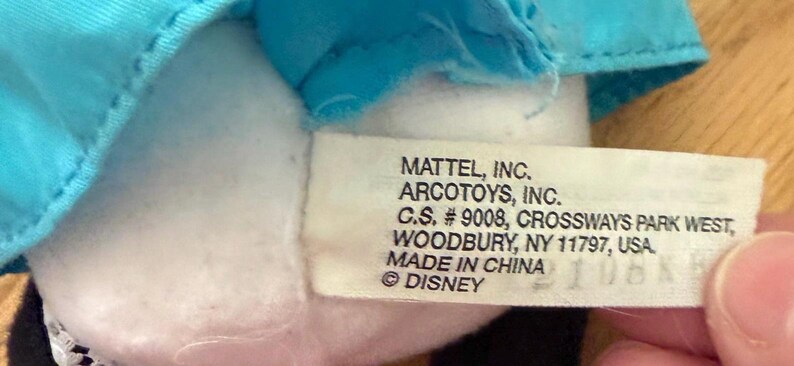 May include: A close-up of a white tag with black text. The tag reads "MATTEL, INC. ARCOTOYS, INC. C.S. #9008, CROSSWAYS PARK WEST, WOODBURY, NY 11797, USA. MADE IN CHINA &copy; DISNEY".